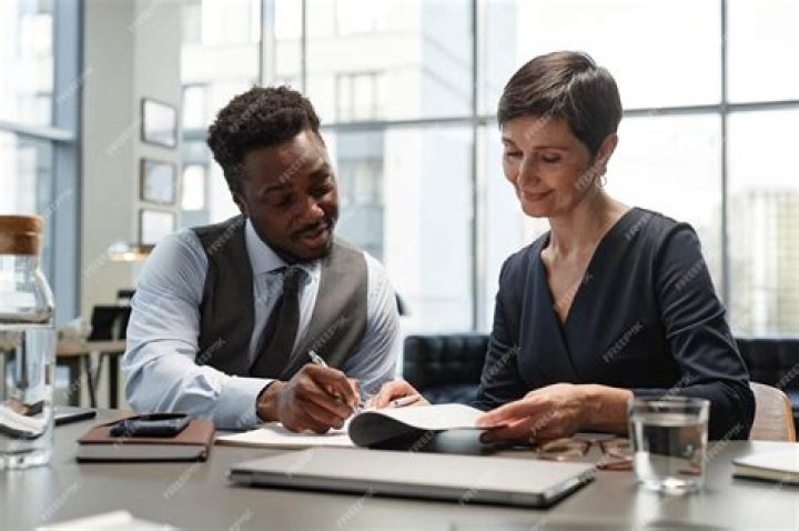 Photo of two individuals looking at laptop screen, other people in background working on laptops